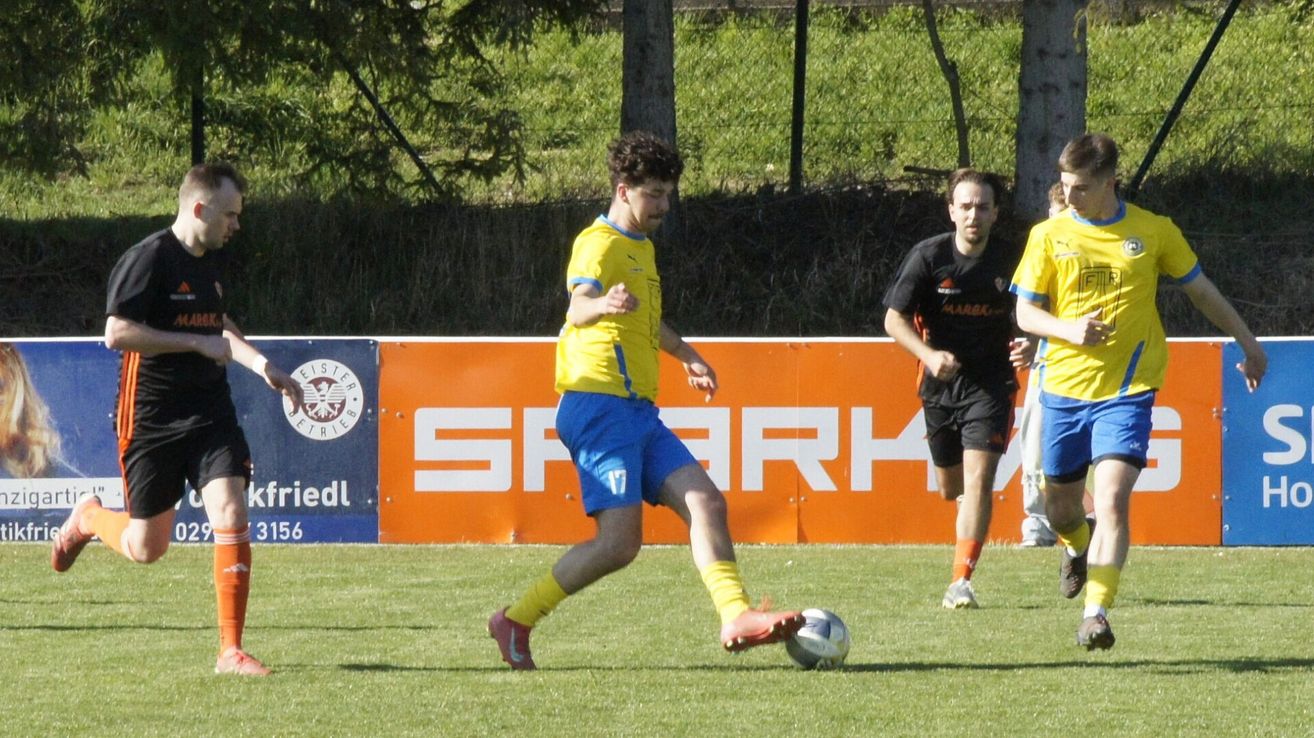 Ein Fußballspiel ist in vollem Gange. Ein Spieler in Gelb jagt dem Ball hinterher, während ein anderer Spieler in Schwarz von rechts kommt. Ein orangefarbener Werbebanner ist im Hintergrund.