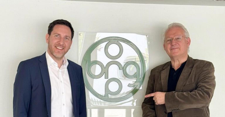 Two men standing next to each other in front of a white wall with a green logo on it. One man is pointing at the logo.