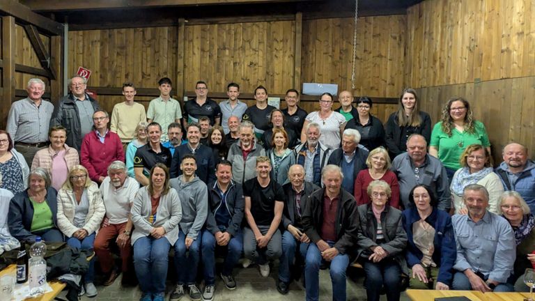 Eine Gruppe von Menschen in unterschiedlicher Kleidung posiert für ein Foto in einem Holzraum. Einige sitzen, andere stehen.