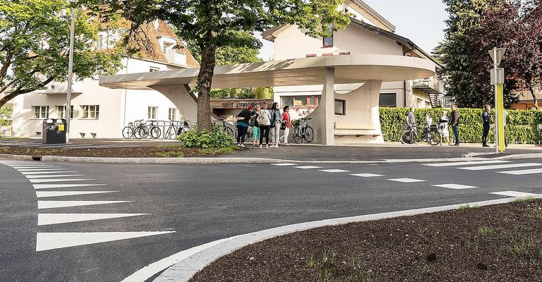 Several people are gathered under a modern, arched bike shelter in a suburban setting. Bicycles are parked nearby, and two individuals are riding bicycles in the background.
