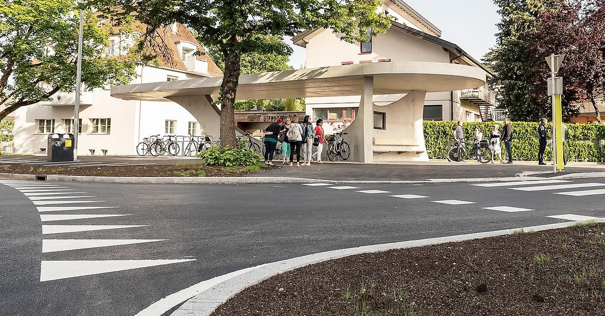 Several people are gathered under a modern, arched bike shelter in a suburban setting. Bicycles are parked nearby, and two individuals are riding bicycles in the background.