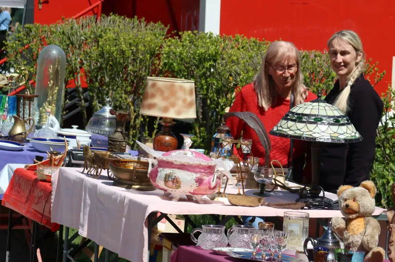 A table covered with various antiques like a teapot, glasses, and lamps, with a woman standing nearby in front of a red building.