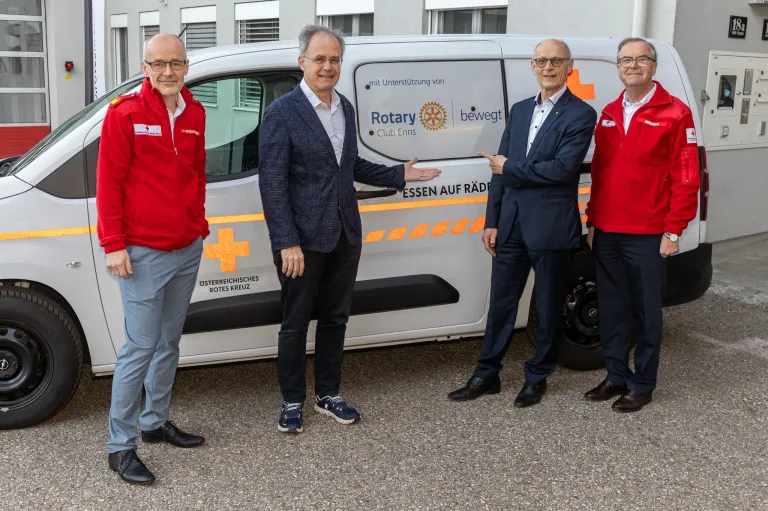 Vier Männer stehen neben einem weißen Van mit 'Rotary' und rotem Kreuz Logo. Sie tragen rote Jacken und formale Kleidung. Der Mann in der Mitte zeigt auf den Van.