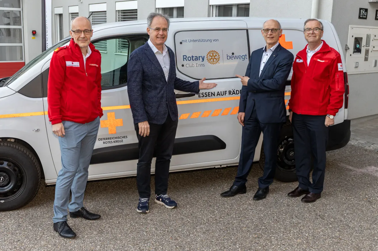 Vier Männer stehen neben einem weißen Van mit 'Rotary' und rotem Kreuz Logo. Sie tragen rote Jacken und formale Kleidung. Der Mann in der Mitte zeigt auf den Van.
