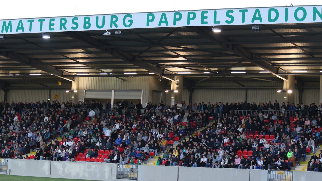 A large crowd fills the Pappelstadion, with many wearing green and white. The stadium's sign reads 'Pappelstadion'. The crowd is spread across red seats, and the stadium has a metal roof.