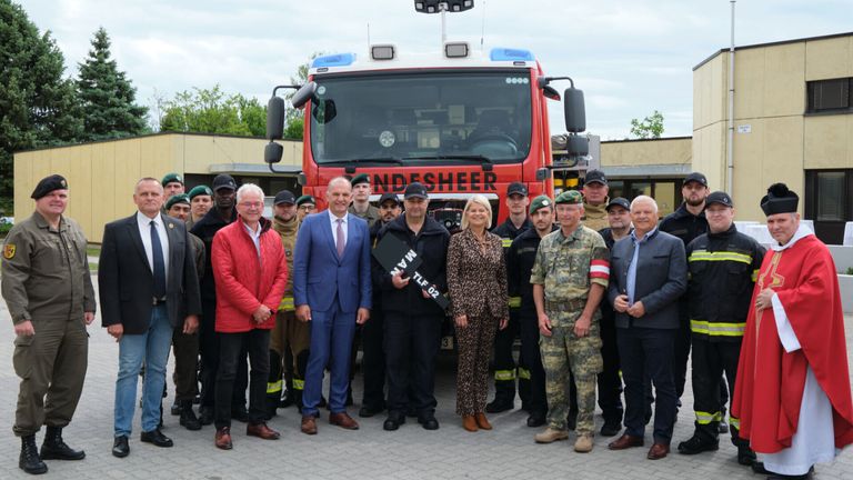 Eine Gruppe von Menschen steht vor einem roten Feuerwehrauto. Ein Mann in Anzug hält eine Plakette. Eine Frau in einem Leoparden-Outfit steht neben ihm.