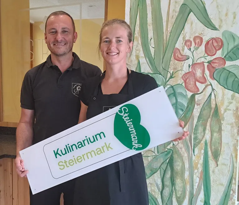 Ein Mann und eine Frau stehen nebeneinander und halten ein weißes Schild mit grünem Text, auf dem 'Kulinairium Steiermark' steht. Sie lächeln und scheinen für ein Foto zu posieren.