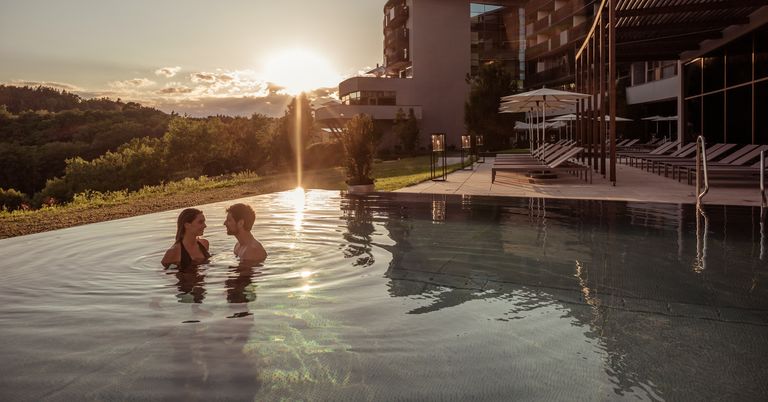 Ein Paar genießt ein Schwimmen in einem Außenpool bei Sonnenuntergang mit einem Gebäude und Liegestühlen im Hintergrund.