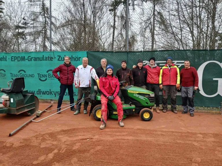 A group of people dressed warmly stand behind a John Deere lawnmower on a dirt field, with a green fence and trees in the background.