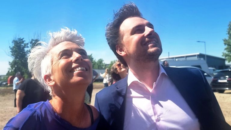 Two people are smiling and standing close together under a blue sky. The woman has white hair and the man has a beard. They are wearing formal clothes.