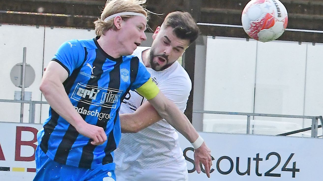 Zwei Fußballer in einem Spiel, einer in einem blau-schwarzen Trikot, der andere in Weiß, beide in Bewegung.