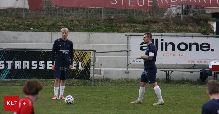 Zwei Fußballspieler in blauen Trikots stehen auf dem Feld. Einer schaut auf den Ball, während der andere ihn ansieht. Ein Schild mit dem Wort 'allin' steht hinter ihnen.