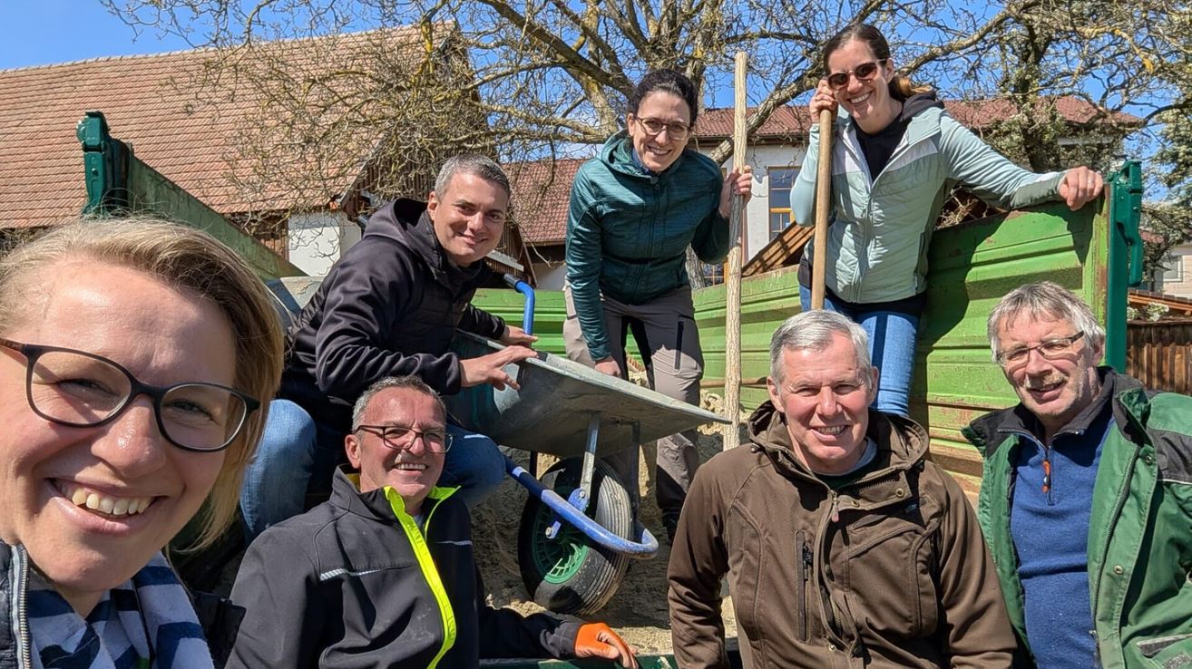 Fünf Menschen lächeln und posieren für ein Foto im Freien. Zwei halten Stöcke, einer einen Schubkarren. Im Hintergrund ist ein Haus und ein Baum zu sehen.