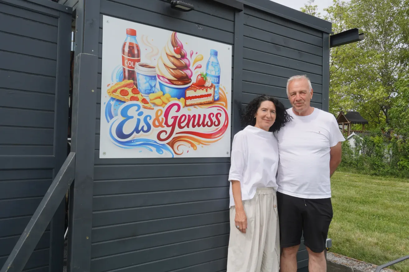 Ein Mann und eine Frau stehen vor einem Stand namens Eis & Genuss mit einem bunten Plakat von Eis, Pizza und Getränken.