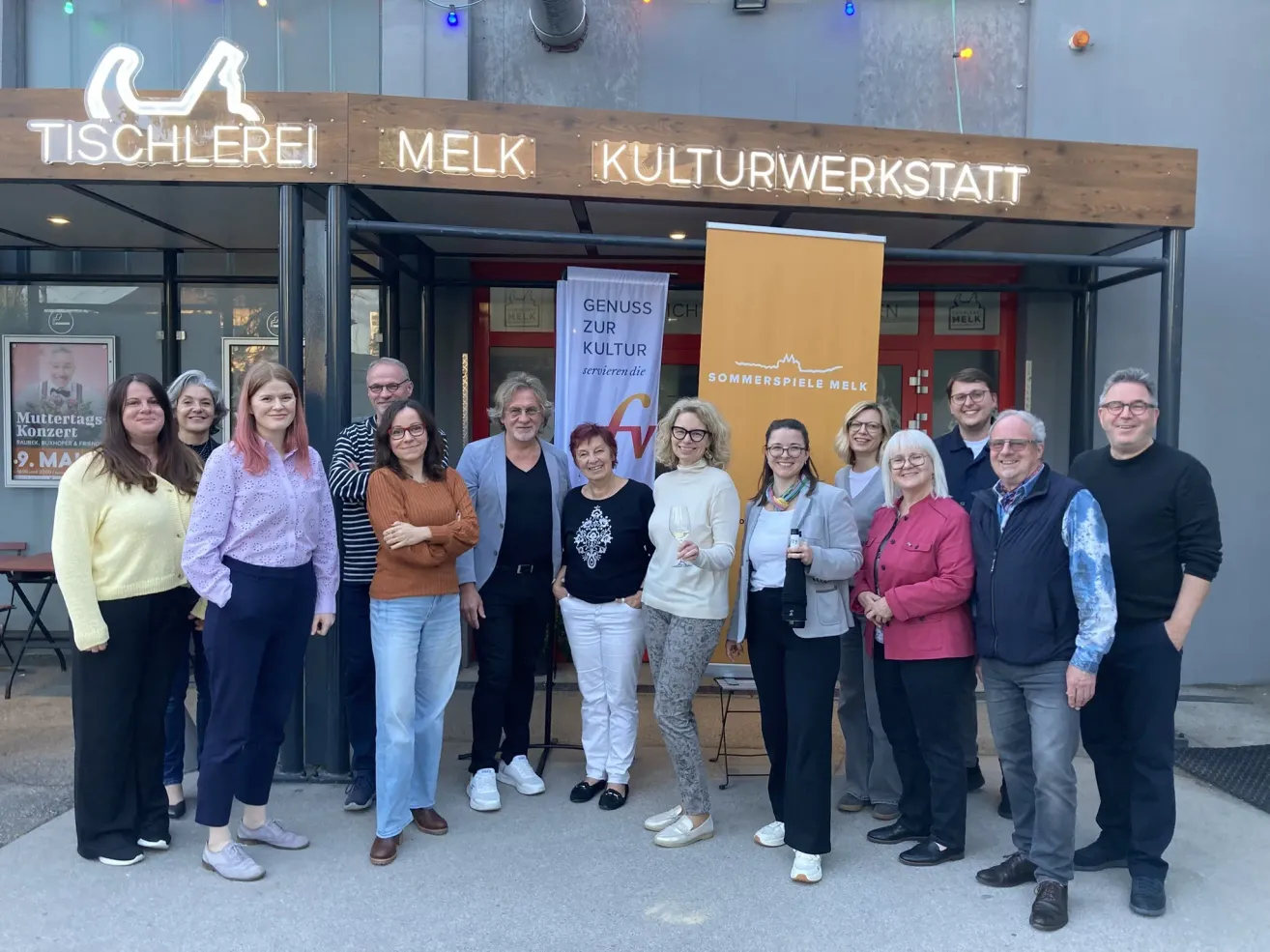 Eine Gruppe von Menschen, sowohl Männer als auch Frauen, stehen vor einem Gebäude mit dem Namen 'Melk Kulturwerkstatt' darauf. Sie lächeln und posieren für ein Foto.