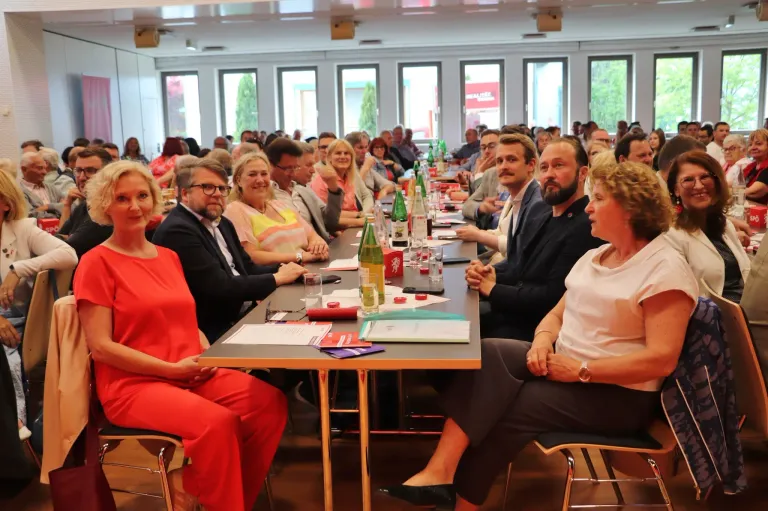 Eine Gruppe von Menschen sitzt um einen langen Tisch in einem Raum. Sie tragen formale Kleidung, und einige tragen Brillen. Auf dem Tisch befinden sich Flaschen, Gläser und andere Gegenstände. Der Raum hat mehrere Fenster mit Jalousien und eine Deckenventilation.