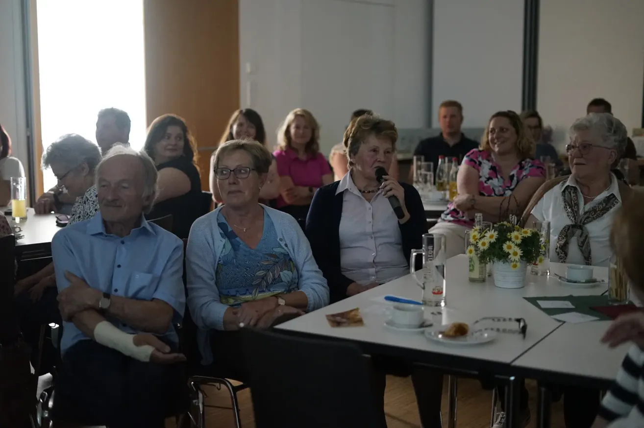 Eine Gruppe von Menschen sitzt in einem Raum. Eine Frau spricht in ein Mikrofon. Ein Tisch mit Blumen und einer Tasse steht davor.