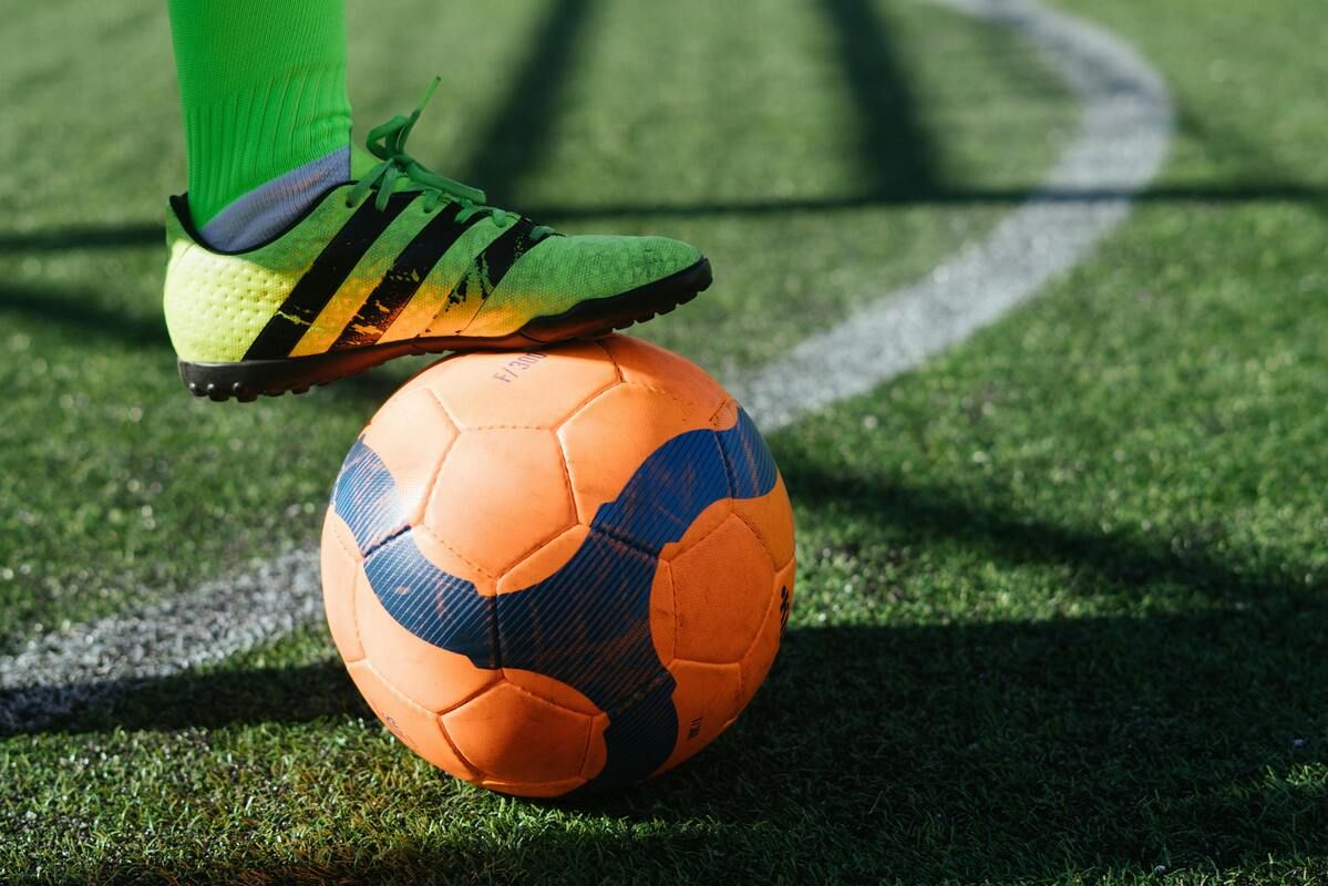 Ein Fußballspieler mit grünen Socken und Adidas-Schuhen steht auf einem Fußball auf einem Rasenfeld. Der Ball hat ein orange-blaues Design.