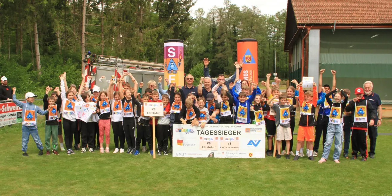 Eine Gruppe von Kindern und Erwachsenen steht auf einem Rasenfeld und hält ein großes Schild. Sie tragen alle einheitliche Uniformen. Sie lächeln und heben ihre Hände. Hinter ihnen stehen zwei Banner und ein Feuerwehrwagen.