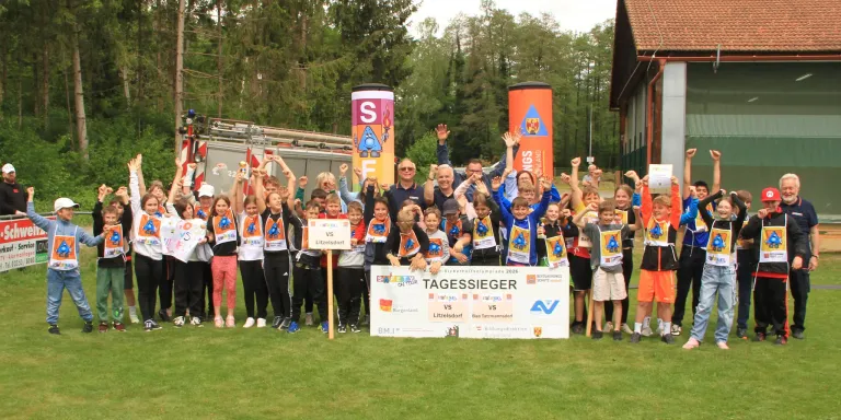 Eine Gruppe von Kindern und Erwachsenen steht auf einem Rasenfeld und hält ein großes Schild. Sie tragen alle einheitliche Uniformen. Sie lächeln und heben ihre Hände. Hinter ihnen stehen zwei Banner und ein Feuerwehrwagen.