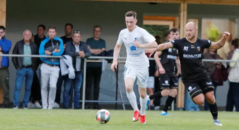 A soccer game in progress with players in white and black uniforms, one player dribbling the ball. Spectators watch from behind a fence.