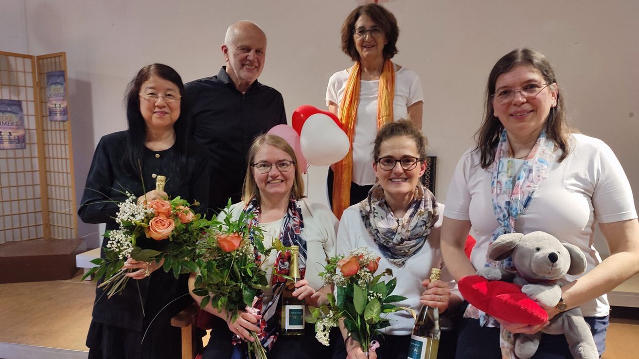 Eine Gruppe lächelnder Personen steht zusammen, hält Blumen und Flaschen und hat Luftballons im Hintergrund.