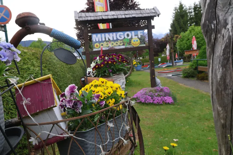 A mini-golf course with flowers and a sign, situated outdoors with a pathway and a small garden.