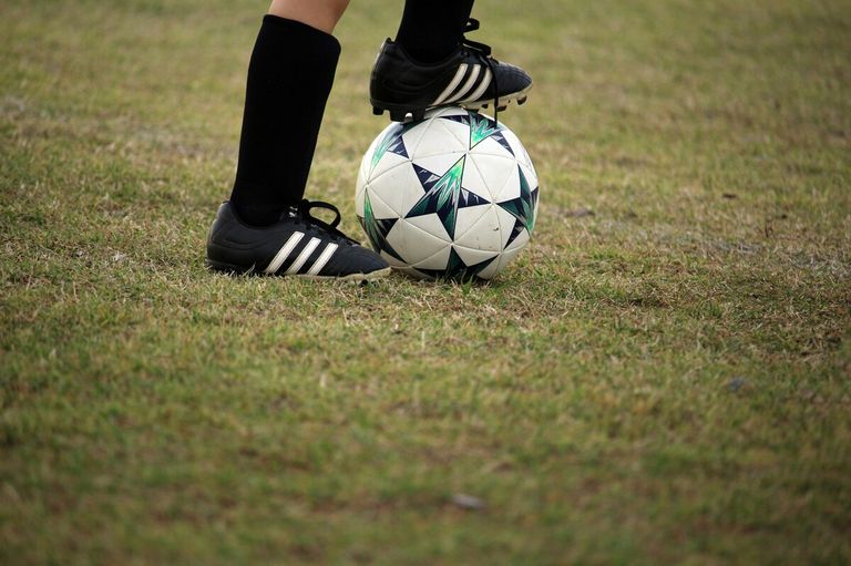 Ein Fußballspieler steht auf einem Fußball mit einem Fuß darauf. Der Spieler trägt schwarze Socken und Adidas Fußballschuhe.