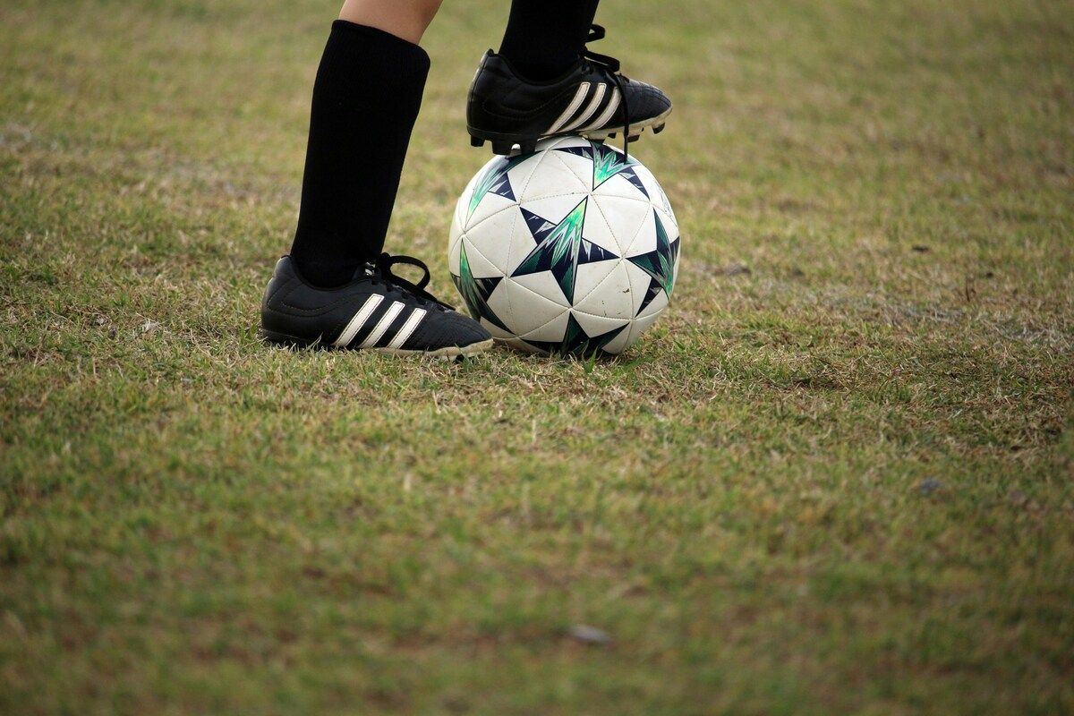 Ein Fußballspieler steht auf einem Fußball mit einem Fuß darauf. Der Spieler trägt schwarze Socken und Adidas Fußballschuhe.