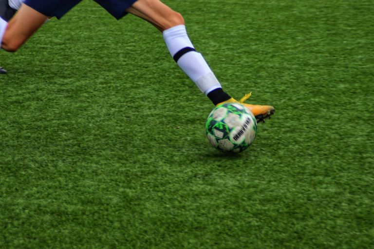 A soccer player kicks a soccer ball on a grassy field. The ball is green and white with a black design.