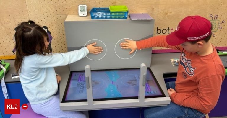 Zwei Kinder spielen in einem Raum mit einer hölzernen Wand mit einem interaktiven Bildschirm. Ein Digitaluhr und einige Aufbewahrungsboxen sind oben.