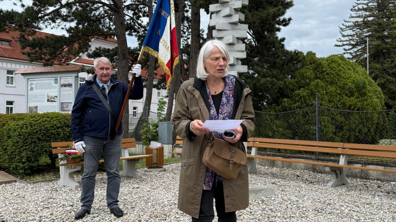An elderly woman stands holding a paper and flag, while a man with a flag stands behind her, both near a bench and a building.