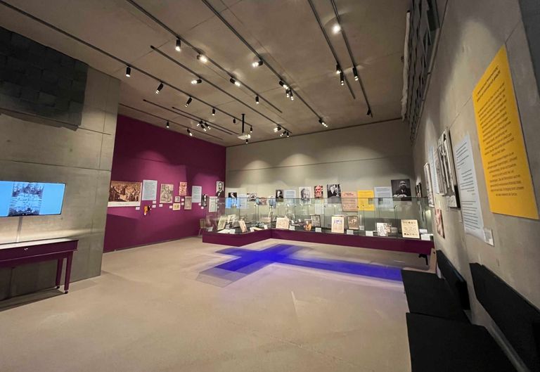 Ein leeres Museum mit einer lila Wand. Verschiedene Gegenstände sind in einer Glasvitrine ausgestellt. An der Wand sind mehrere Papiere befestigt. Die Decke hat Beleuchtung.