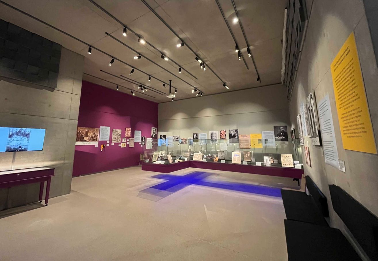 Ein leeres Museum mit einer lila Wand. Verschiedene Gegenstände sind in einer Glasvitrine ausgestellt. An der Wand sind mehrere Papiere befestigt. Die Decke hat Beleuchtung.