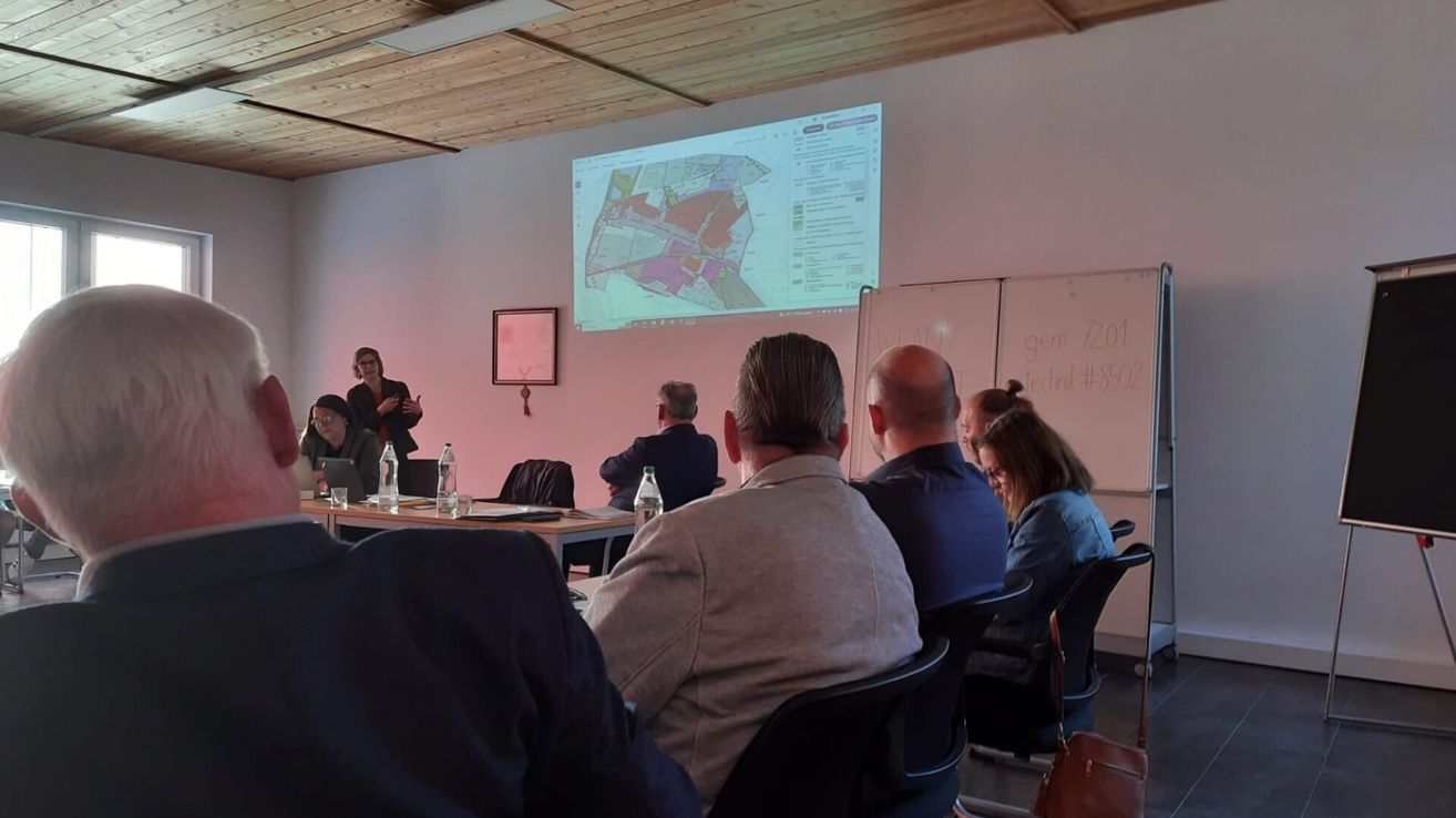 Eine Gruppe von Menschen sitzt in einem Raum und beobachtet eine Präsentation an einer Projektionsleinwand. Eine Frau steht neben einer Tafel, und eine Karte wird auf dem Bildschirm angezeigt.