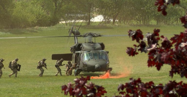 Ein Militärhubschrauber mit Soldaten, die aussteigen, einer hält einen Hund, auf einem Grasfeld mit rotem Rauch am Boden.