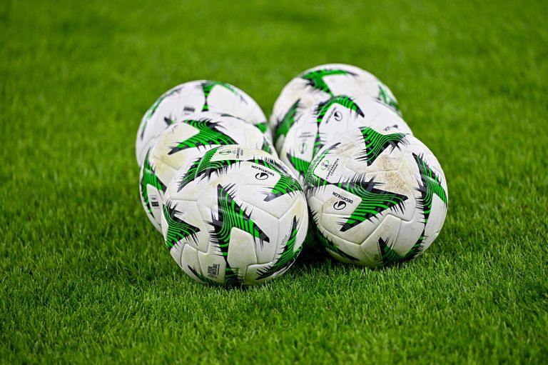 Five soccer balls with green and black designs are on a grass field. They appear to be used for a game.