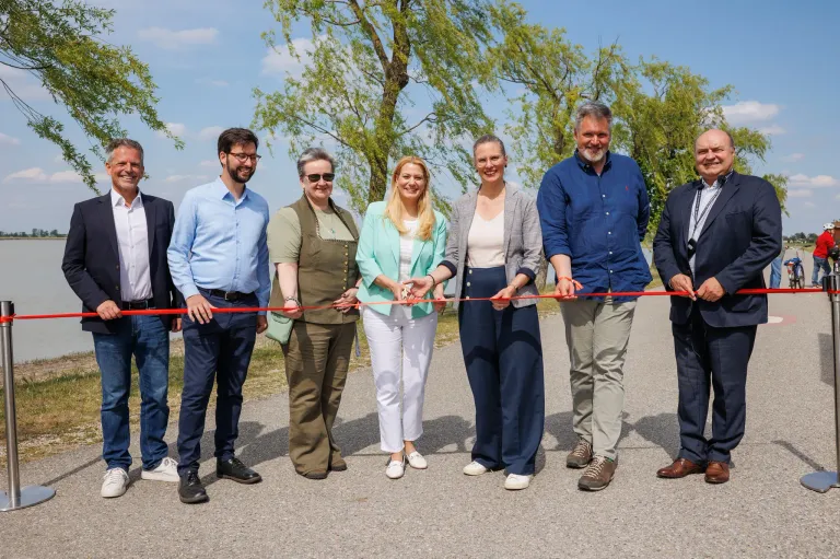 Eine Gruppe von Menschen steht um ein rotes Band und lächelt, während sie es halten. Sie scheinen ein Eröffnungsereignis zu feiern. Im Hintergrund sind Bäume zu sehen.