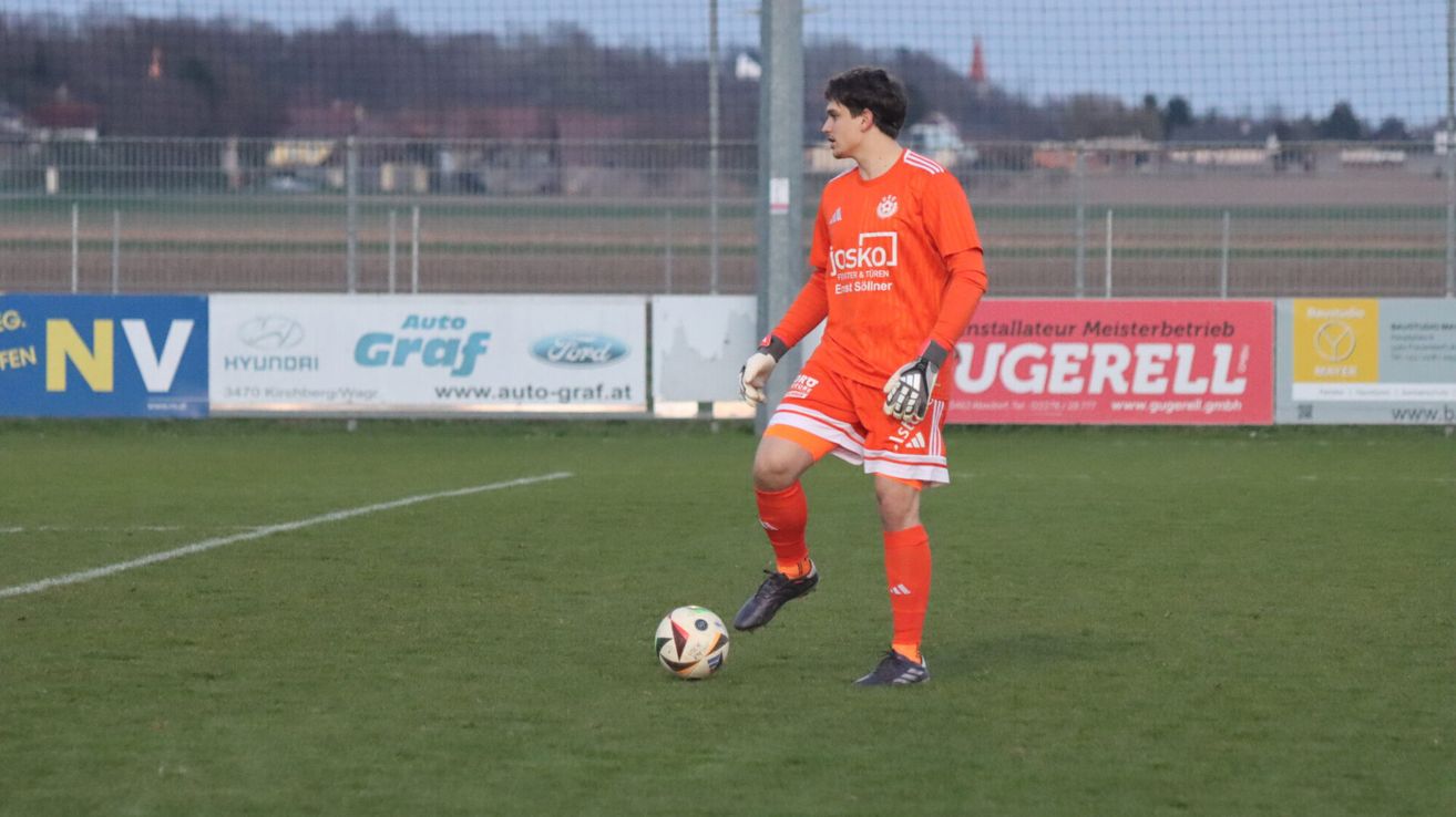 Ein Fußballspieler in orangem Trikot ist dabei, einen Ball auf einem Rasenfeld zu schießen. Der Spieler trägt Handschuhe und Schuhe. Dahinter ist ein verschwommener Hintergrund mit einem Zaun, einem Banner und einem Berg.