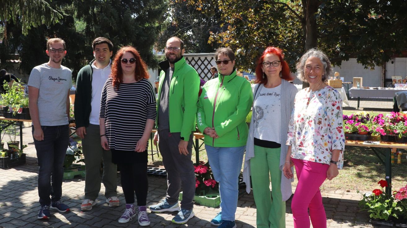 Eine Gruppe von Menschen in grüner und lässiger Kleidung steht in einem Park und lächelt für ein Foto. Sie stehen in der Nähe eines Baumes, eines Holztisches und eines Topfes.