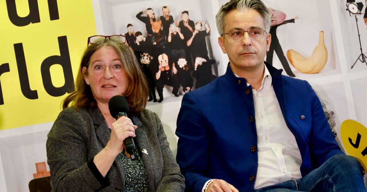 A woman with glasses and a microphone speaks while a man in glasses and a blue jacket listens. Behind them is a poster of people in costumes.