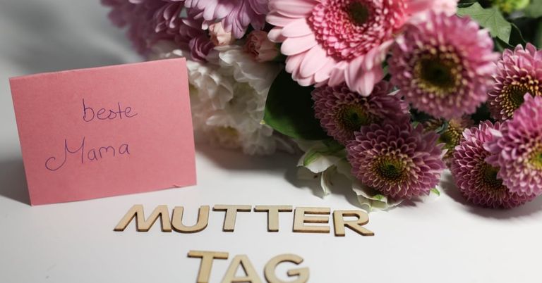 Eine rosa Karte mit dem Wort 'Mutter' darauf, neben einem Strauß aus rosa und weißen Blumen.