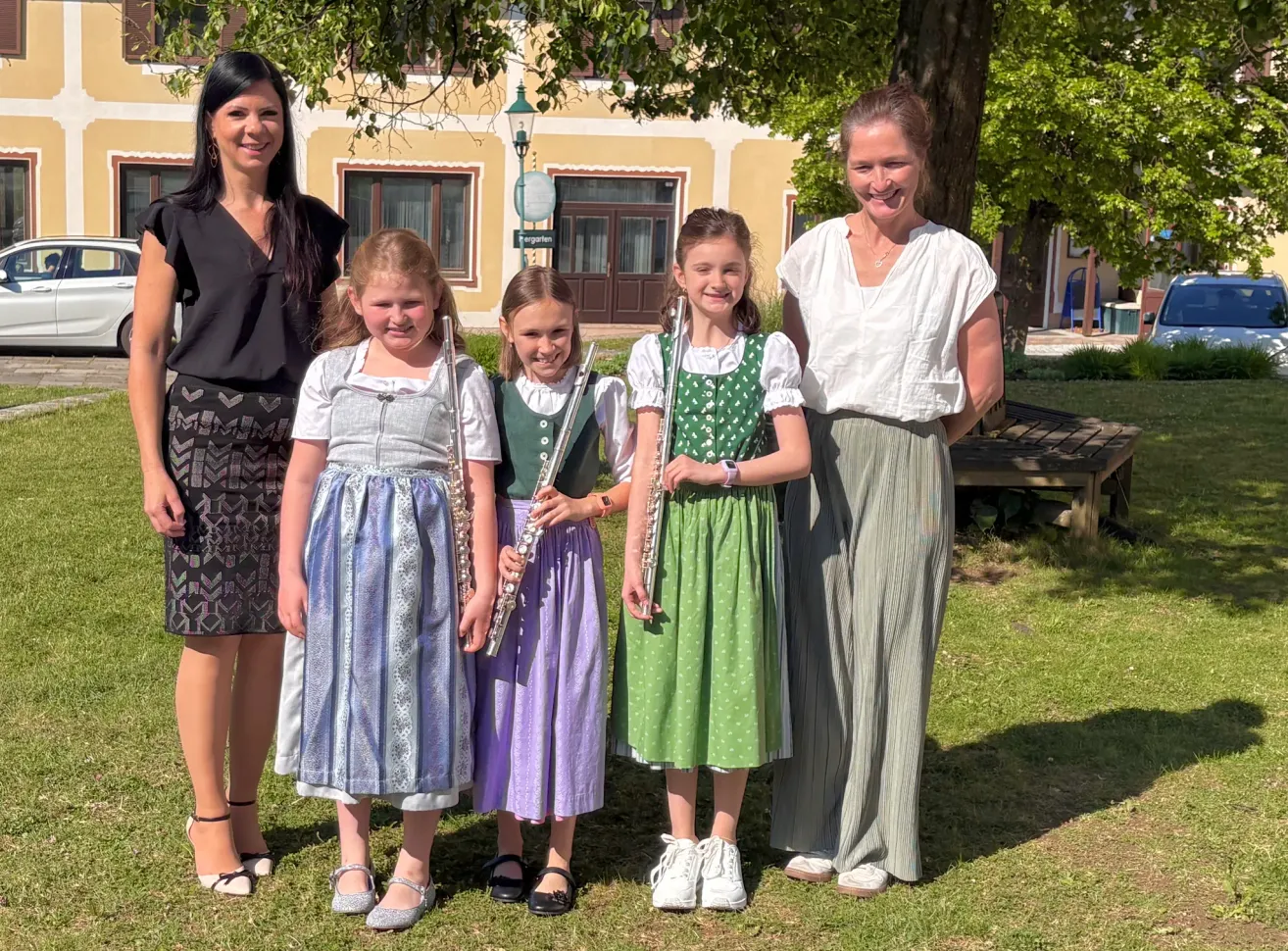 Drei junge Mädchen in traditionellen Kostümen posieren mit ihren Flöten für ein Foto. Zwei Frauen im Hintergrund lächeln für die Kamera. Ein Gebäude mit Fenstern und einer Straßenlaterne ist im Hintergrund.