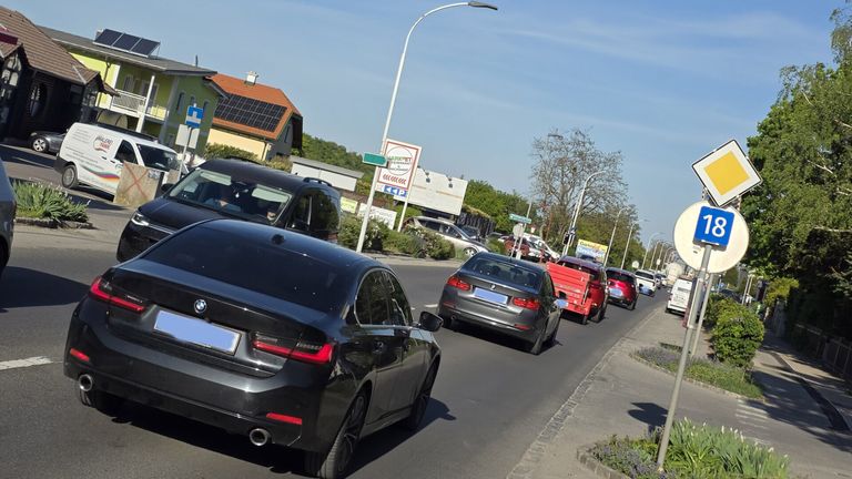 A street with several cars, including a black BMW and a gray BMW, is lined with streetlights. A house with solar panels on the roof is on the left.