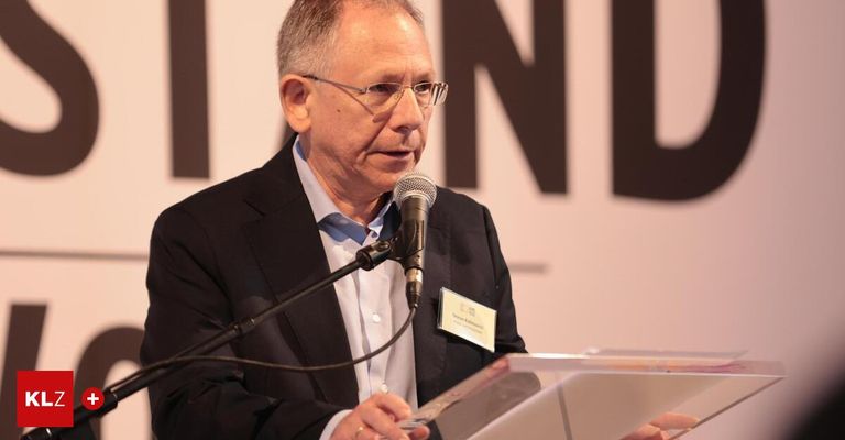 Ein älterer Mann mit Brille steht an einem Rednerpult und spricht in ein Mikrofon. Er trägt einen schwarzen Anzug und ein blaues Hemd. Eine Namensplakette mit Text ist an seinem Jackett befestigt.