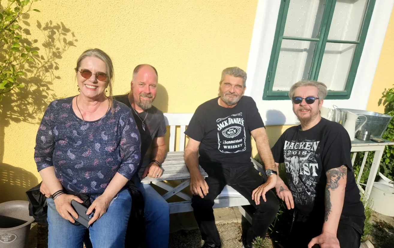 Vier Menschen sitzen auf einer Bank draußen. Die Frau trägt ein Blumenhemd und lächelt. Die Männer tragen schwarze T-Shirts. Einer hat 'Jack Daniel's' auf seinem Shirt. Die Wand hinter ihnen ist gelb.