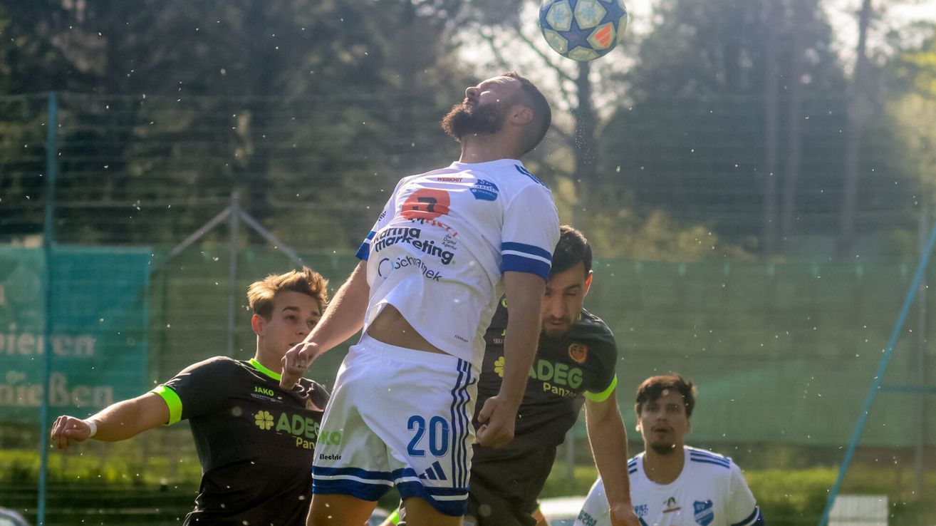 Ein Fußballspieler mit der Nummer 20 auf seinem weißen Trikot springt auf, um den Ball zu köpfen, während seine Teamkollegen versuchen, ihn zu blockieren.