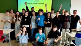 Schüler posieren für ein Foto in einem Klassenzimmer mit einer Projektionsleinwand, auf der FiSci im Hintergrund zu sehen ist. Sie alle lächeln und zeigen Daumen hoch.
