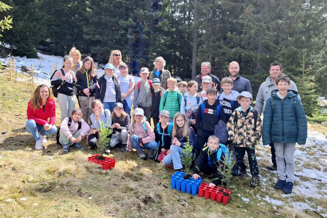 Eine Gruppe von Menschen in einem bewaldeten Gebiet, Erwachsene und Kinder zusammen, lächeln für ein Foto. Einige Kinder halten Setzlinge. Der Hintergrund zeigt Bäume und eine verschneite Landschaft.