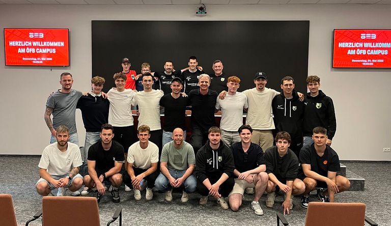 Eine Gruppe von Männern in Sportkleidung posiert für ein Foto in einem Raum mit einer Tafel, einige sitzend und andere stehend.